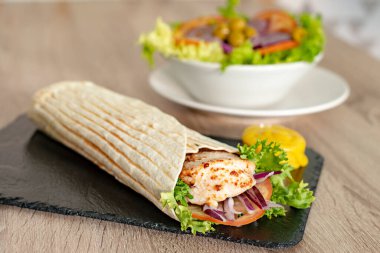 Kızarmış tavuk dürümü, kırmızı soğanlı domates dilimleri, marul ve hardal sosu ahşap masada.