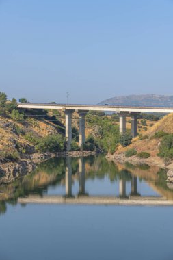 Nehir üzerindeki bir yol köprüsü, yaz günü platformda yol trafiği varken su yüzeyindeki direklerin yansımasını yansıtıyor.