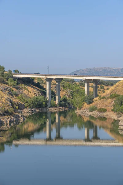Nehir üzerindeki bir yol köprüsü, yaz günü platformda yol trafiği varken su yüzeyindeki direklerin yansımasını yansıtıyor.