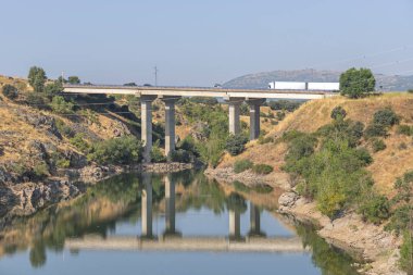Nehir üzerindeki bir otoyol köprüsü, su yüzeyindeki direklerin yansımasını ve güvertedeki yol trafiğini yansıtıyor.