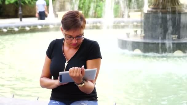 Νεαρή γυναίκα με τον υπολογιστή tablet που κάθεται στο πάρκο της πόλης — Αρχείο Βίντεο