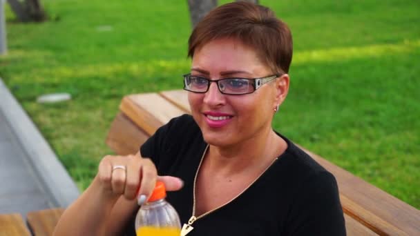 Cute young woman drinking fresh lemonade on bench in the park — Stock Video