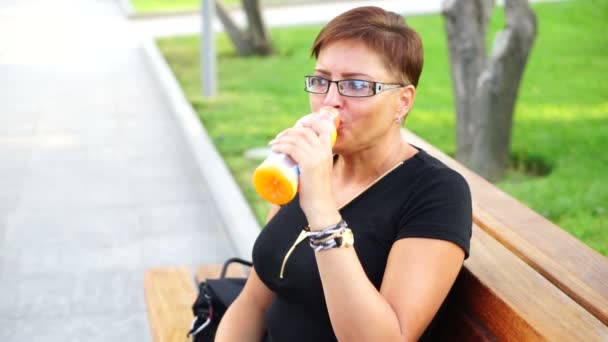 Cute young woman drinking fresh lemonade on bench in the park — Stock Video