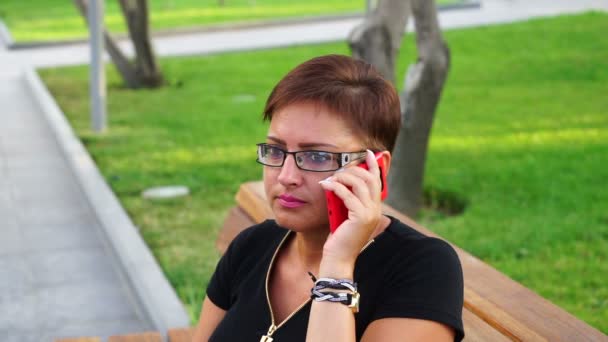 Mujer joven en gafas hablando en el teléfono móvil en un parque de la ciudad . — Vídeos de Stock