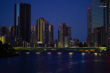 Gökdelenlerin aydınlattığı ve derin mavi bir alacakaranlık altında nehirde yansıyan gökdelenlerin olduğu bir şehrin gece manzarası.