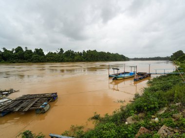Yoğun bulutlu bir gökyüzünün altındaki bulanık, kahverengi bir nehrin geniş bir manzarası, yemyeşil tropikal bir kıyı şeridine kenetlenmiş küçük kayıklar ve ahşap sallar..