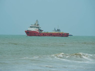 Kırmızı ve beyaz bir endüstriyel tedarik gemisi ufuktaki engin, puslu gökyüzünün altında dalgalı turkuaz deniz suyunda yol alır..