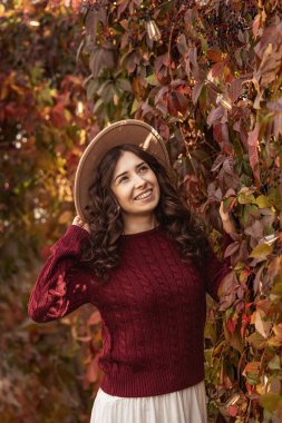 Şapkalı ve kırmızı kazaklı güzel bir kadın renkli yaprakların yanında sonbahar gününün tadını çıkarıyor. Sıcak ve samimi sonbahar portresi, mevsimlik moda ve yaşam tarzı konsepti