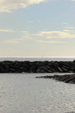 Önünde koyu kayalık bir dalgakıran ve ufka uzanan sakin gümüş bir deniz bulunan dikey kıyı manzarası..