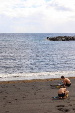 Okyanus kıyısındaki siyah bir kumsalda renkli kum oyuncaklarıyla oynayan iki çocuğun olduğu dikey sahneler..