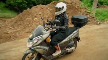 Genç bir kadın, çamurlu bir yolda motorsikletle ormanın içinden su birikintisine doğru gidiyor.