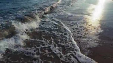 Yumuşak okyanus dalgaları, parıldayan güneş ışığı yansımalarıyla kumlu kıyılarda yuvarlanarak yumuşak köpük dokusu ritmik hareketi ve sakin kıyı güzelliğini canlı detaylarıyla yakalıyor. Yüksek kalite 4K görüntü.
