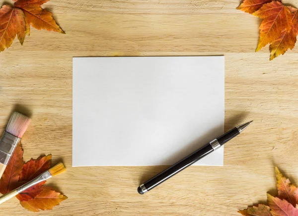 Background texture with wooden table and yellow  and orange autumnal leaves. Frame, made from pen, painting brushes, autumn leaves and white paper — Stock Photo, Image