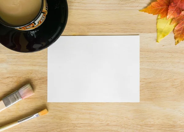 Painting brushes, autumn leaves, black cup of coffee and white paper on wooden table — Stock Photo, Image