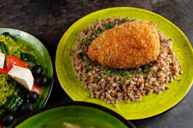 Bir tabak kızarmış tavuk, otlarla süslenmiş bir karabuğday yığınının üzerinde duruyor. Yemeğe marul, salatalık, domates, siyah zeytin ve peynirden yapılmış taze bir salata eşlik eder..