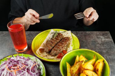 Bir insan kızarmış kebap yemeye hazırlanıyor. Bir tabak dolusu ekmek ile servis ediliyor. Salata ve kızarmış patates de var. Bir bardak içecek yemeğe ilave eder..
