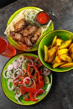 Tabaklar renkli tabaklarda servis edilir, soslu ızgara et, taze sebze salatası ve kırmızı içeceğin yanında kızarmış patates gösterilir. Manzara canlı ve parlak..