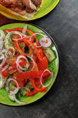 Yeşil bir tabağa kesilmiş salatalık, domates, kırmızı biber ve soğanlı taze bir salata yerleştirilir. Arka planda bir et yemeği görülüyor.