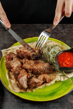 Bir insan, yanında sebze ve sos ile servis edilen ızgara et tabağını keser. Yemek rengarenk bir tabağa konur..