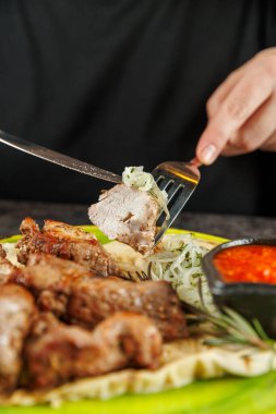 Taze otlar ve baharatlı soslar yanına yerleştirilirken bir kişi ızgara eti kesmek için çatal ve bıçak kullanır. Ayar gündelik ve davetkar..