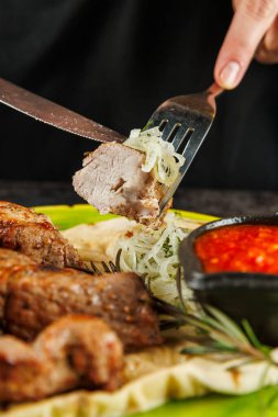 Birisi yanında soğan dilimleri tutarken ızgara et dilimini çatalla keser. Yanında bir kase baharatlı sos var. Bu sahne geleneksel yemek düzenini gösteriyor..