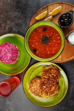 Tabaklar arasında bir kase kırmızı çorba, yeşil krep ve limon dilimleri ve ahşap tabakta zeytinlerle düzenlenmiş pembe salata yer alıyor. Bir içki de görünür..