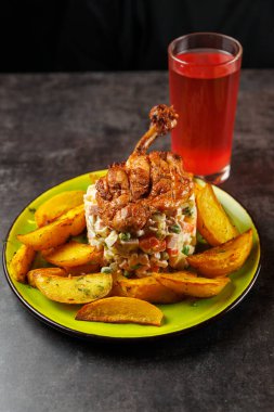 Tabakta sebze salatasının üzerinde kızarmış tavuk ve yanında patates dilimleri yer alıyor. Koyu bir masada, tabağın yanında bir bardak kırmızı içki duruyor..