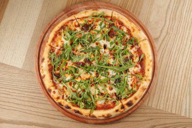 Taze pizza ahşap bir tahtanın üzerinde duruyor. Üstünde bir sürü roka var. Arka plan basit bir tahta masayı gösteriyor. Bu sahne arkadaşlarla yemek yemeye ve eğlenmeye davet ediyor..