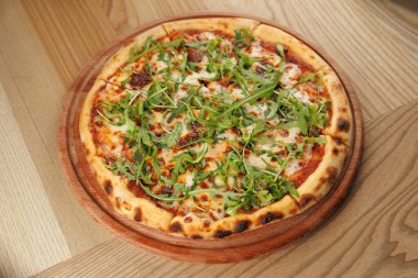 Yuvarlak bir pizza ahşap bir tabağa konur. Tabakta erimiş peynir, domates sosu ve taze roka yer alıyor. Sıradan bir restoran ortamında hafif bir ahşap masaya kurulur..