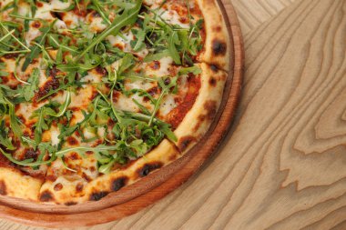 Lezzetli pizza, eritilmiş peynir ve taze roka ile kaplı ahşap bir tabakta duruyor. Sıradan yemek atmosferi bu yemeğin tadını çıkarmak için mükemmel bir ortam hazırlıyor..