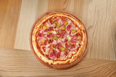 Yuvarlak bir pizza ahşap bir yüzeye yerleştirilir. Pizza üzerinde jambon, yeşil biber ve zeytin dilimleri var. Tadını çıkarmaya hazır..
