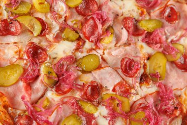 Bir tabak, üzerinde jambon, turşu ve soğan bulunan yeni pişmiş bir pizza içeriyor. Renkli malzemeler, sıradan bir yemek ortamında altın bir kabuk üzerinde sergileniyor..