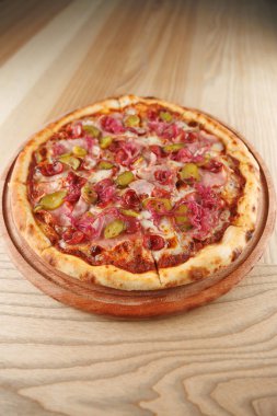 Yeni pişmiş bir pizza yuvarlak ahşap bir tabakta oturur. Pizzada çeşitli et ve sebzeler, erimiş peynir ve kabuğun üzerinde görünen baharatlar yer alıyor..