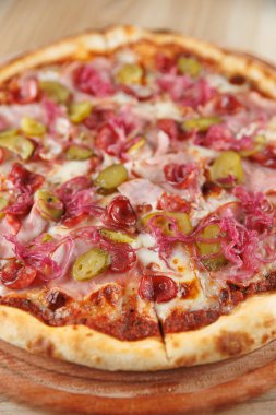 Yuvarlak bir pizza taze pişmiş ve dilimlenmiş jambon, turşu ve kırmızı soğanla kaplıdır. Tabak ahşap bir tabakta sunuluyor, malzemelerin canlı renklerini vurguluyor..