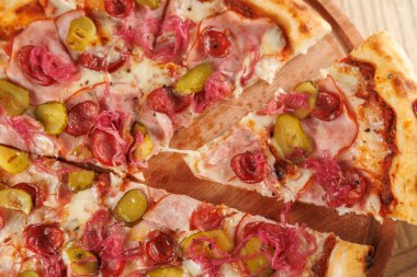 Dilimlenmiş pizza, içinde jambon, pepperoni, yeşil zeytin ve soğan gibi malzemeler bulunan ahşap bir tahtaya konur. Bir parça çıkartılır, kalitesiz dokusu ve canlı renkleri sergilenir..