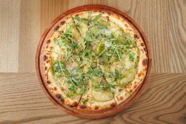 Yuvarlak pizza, taze yeşil malzemeler ve patates dilimleriyle harmanlanmış dokular sergiliyor. Bir restoranın ahşap tahtasında sunuluyor..