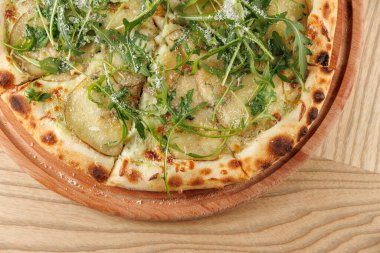 Yuvarlak ahşap bir tabakta pizza servis edilir. Sergide dilimlenmiş patates, taze yeşillik ve peynir gibi malzemeler yer alıyor. Dekor, ahşap yüzeyi olan sıcak bir yemek ortamı gösteriyor..