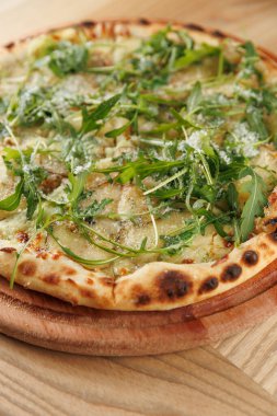 Yuvarlak bir pizza, taze roka ve peynir serpiştirilmiş ahşap bir tahtanın üzerinde duruyor. Kabuk çıtır çıtır görünüyor ve malzemeler de yemek için taze bir görünüm sağlıyor..