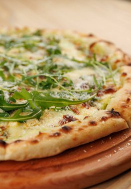 Yuvarlak bir pizza tahta bir tahtaya yerleştirilir. Pizzanın altın bir kabuğu var ve üzeri taze roka ve rendelenmiş peynirle kaplı. Lezzet ve dokuların karışımını sergiliyor..