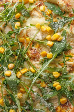 Pizza tavuk, mısır ve taze roka ile hazırlanıyor. Hamur altın ve kıtır kıtır, içeriği ise canlı. Arka planda hareketli bir mutfak var.