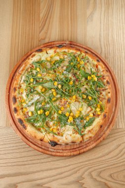 Arugula ve mısırla kaplı yuvarlak bir pizza tahtadan bir pizza tahtasında duruyor. Kabuğu altın kahverengidir, ve taze malzemeler zevk için hazırlanan yemeği vurgular..