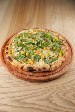 Kıtır kenarlı yuvarlak bir pizza, ahşap servis tabağında sunulan taze ıspanak, mısır ve peynirle kaplıdır. Ayar hafif ahşap bir masa içeriyor..