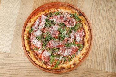 Ahşap tabakta yuvarlak bir pizza, üzerinde pişirilmiş et, taze sebze ve rendelenmiş peynir bulunan bir hamur tabanı içerir. Tabak, gevşek bir lokantada, ahşap bir masaya konur..