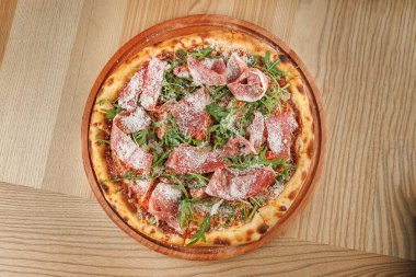 Taze pişmiş pizza, domates sosu, roka ve ahşap bir tabakta peynirle süslenmiş kesilmiş et dilimleriyle kaplı bir kabuk içerir..