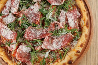 Bir dilim prosciutto, roka ve rendelenmiş peynirle kaplı taze bir pizza ahşap bir tahtaya yerleştirilir. Kabuğu altın gibi ve çıtır çıtır, servise hazır..