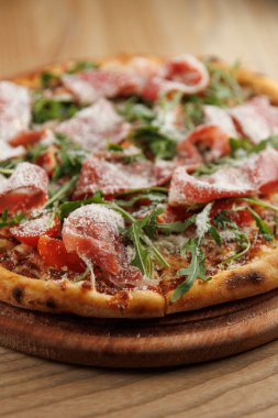 Lezzetli bir pizza ahşap bir tahtada sergilenir. İçinde taze roka, bir dilim prosciutto ve bir tutam peynir var. Bu yemek, sıcak bir yemek atmosferinde keyiflenmeye hazır..