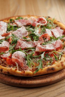 Bir pizza ahşap bir tahtaya yerleştirilir. İçinde domates, roka, jambon ve üzerine peynir serpiştirilmiş bir kabuk bulunur. Ortam sıcak ve davetkar bir ortam sunuyor..