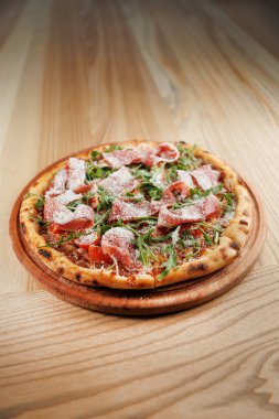 Yuvarlak pizza, domates soslu ince bir kabuk, pişirilmiş et dilimleri ve taze roka içerir. Tahta servis tahtasının üzerinde duruyor, canlı renklerini ve dokularını sergiliyor..