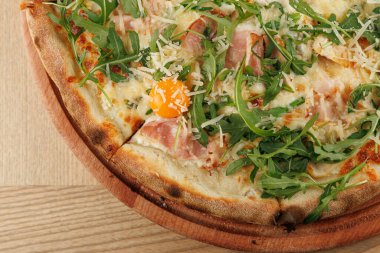 Taze roka, jambon ve çiğ yumurta içeren lezzetli bir pizzanın yakın çekim görüntüsü. Pizza ahşap bir tahtada sergileniyor, çıtır çıtır kabuğu ve malzemelerini sergiliyor..