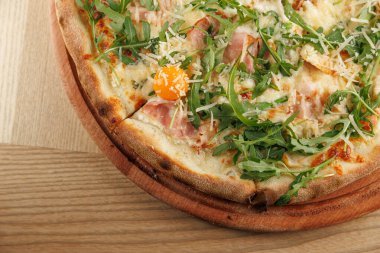 Yeni pişmiş bir pizza ahşap bir tahtanın üzerinde, roka, jambon, peynir ve parlak yumurta sarısı ile kaplıdır. Sahne öğle yemeği sırasında bir kafenin davetkar atmosferini yakalar..